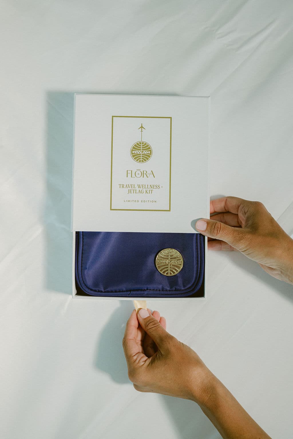 A person's hands are shown opening a white box labeled 'FLORA TRAVEL WELLNESS + JETLAG KIT' to reveal a navy blue satin pouch with a gold emblem inside, set against a white fabric background.