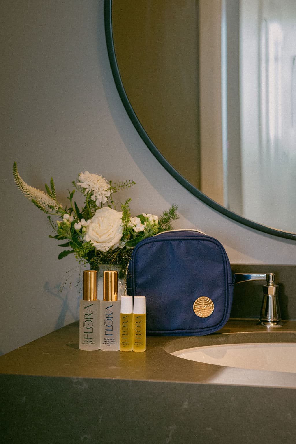 A navy blue toiletry bag with a gold circular logo sits on a bathroom counter next to a mirror, with two gold-capped and white-capped skincare bottles and a floral arrangement in front of it.