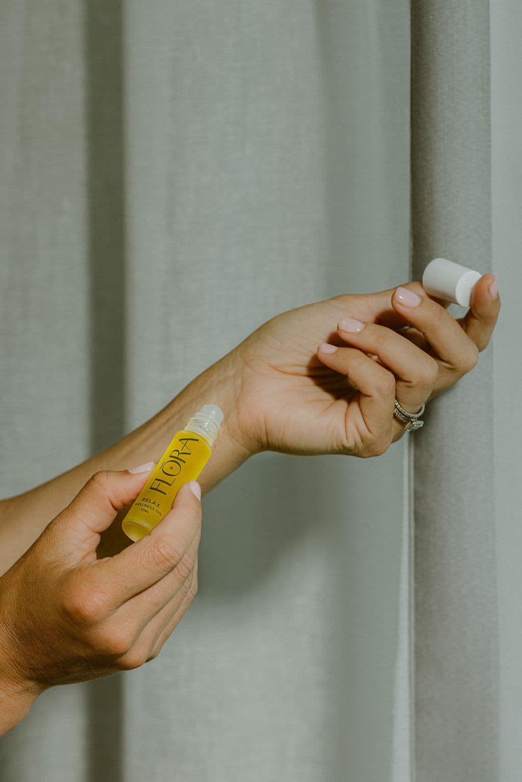 A person applies a small yellow roll-on bottle to their wrist, with the white cap held in the other hand against a soft gray curtain background.