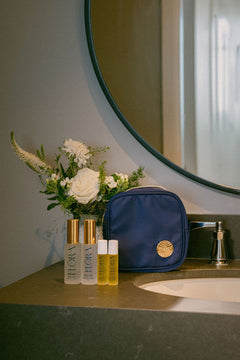 A navy blue toiletry bag with a gold circular logo sits on a bathroom counter next to a mirror, with two gold-capped and white-capped skincare bottles and a floral arrangement in front of it.