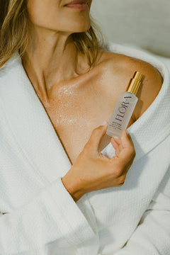 A woman in a white robe holds a frosted bottle labeled FLORA with a gold cap, applying a glossy, shimmering skincare product to her chest.