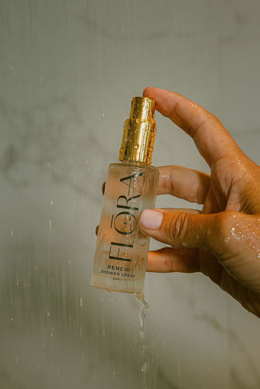 A hand holding a clear shower spray bottle with a gold pump cap under running water, with the brand name FLORA visible on the bottle.