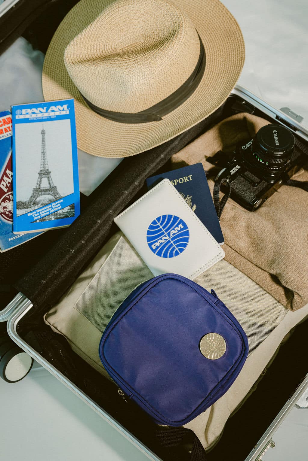 A top-down view of a packed suitcase containing a beige straw hat with a black band, a blue passport, a white passport cover with a blue circular logo, a black camera, a beige towel, and a blue zippered pouch with a silver button.