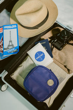 A top-down view of a packed suitcase containing a beige straw hat with a black band, a blue passport, a white passport cover with a blue circular logo, a black camera, a beige towel, and a blue zippered pouch with a silver button.