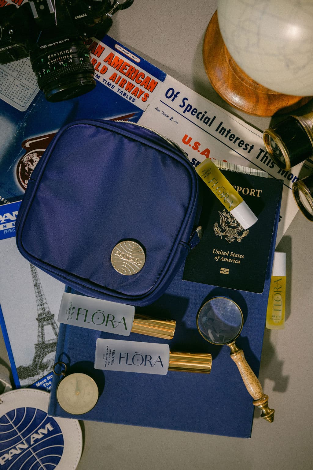 A navy blue zippered travel pouch with a gold coin emblem, styled on top of a blue travel-themed flat lay with a passport, camera, globe, magnifying glass, and other travel accessories.